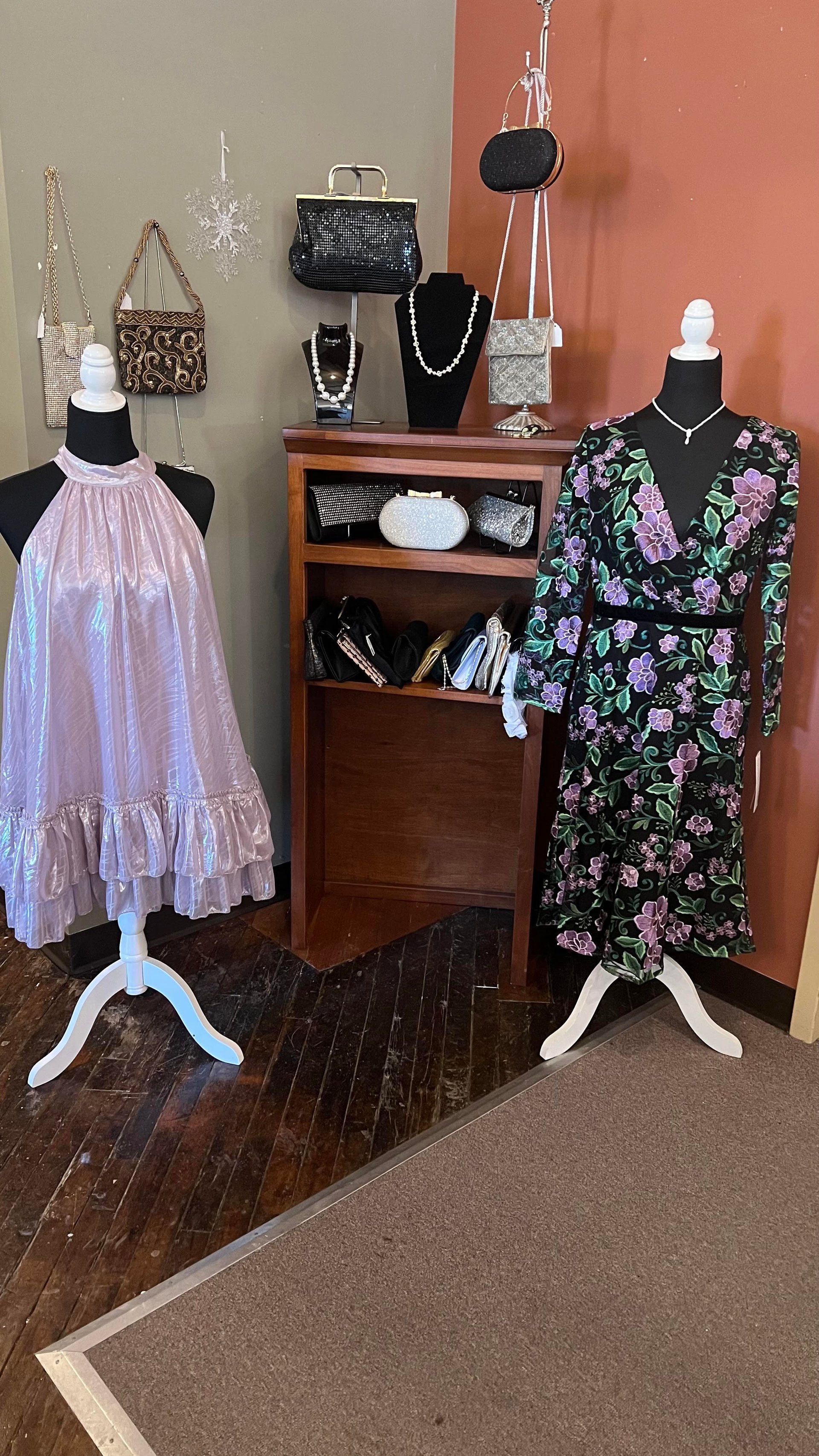 Retail display featuring two mannequins dressed in pink tulle and floral print dresses, with a wooden shelf displaying handbags, jewelry, and accessories against a neutral and coral wall backdrop.