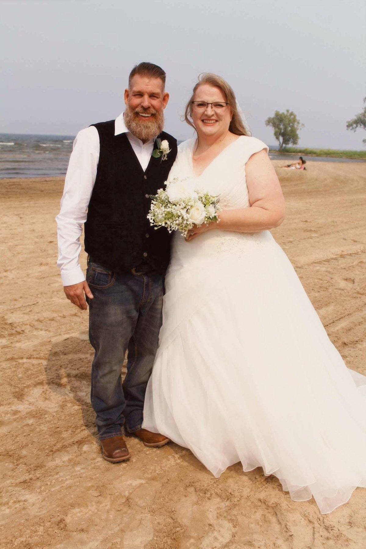 Bride and groom posing together on a beach in wedding attire, holding a white flower bouquet